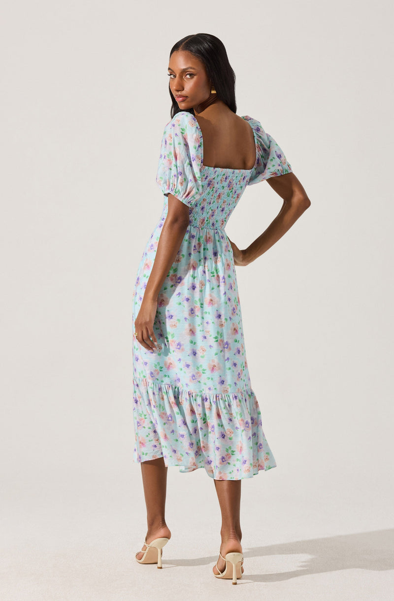 A woman is wearing a light purple midi dress with a floral pattern in pastel colors. The dress has puffed short sleeves, a fitted bodice with a square neckline, and a ruffled hem at the bottom.