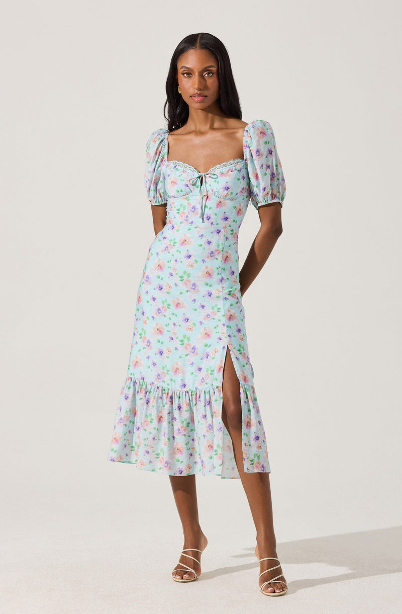 A woman is wearing a midi dress with a pastel floral print. The dress has puffed short sleeves, a square neckline with a lace trim, and a front tie detail at the bust. It features a high slit on one side and a ruffled hem at the bottom, adding a feminine touch. The fabric appears lightweight and flowy. She is paired with strappy high-heeled sandals.