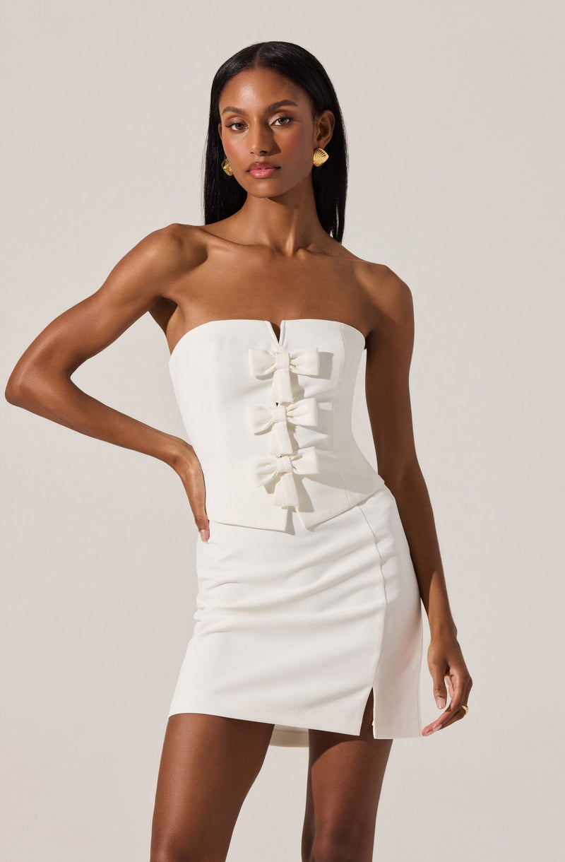 A woman is wearing a strapless white dress with a fitted bodice featuring vertical seams and four decorative bows down the front. The dress is knee-length with a small front slit. She accessorizes with gold earrings and has long, straight black hair.