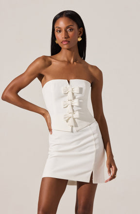A woman is wearing a strapless white dress with a fitted bodice featuring vertical seams and four decorative bows down the front. The dress is knee-length with a small front slit. She accessorizes with gold earrings and has long, straight black hair.