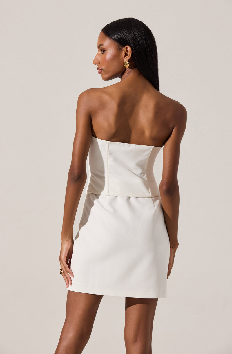 A woman is wearing a strapless, off-white satin dress with a fitted bodice and a slightly A-line skirt. The dress appears elegant and minimalistic, with clean lines and a smooth texture. She accessorizes with gold hoop earrings and a ring on her right hand.