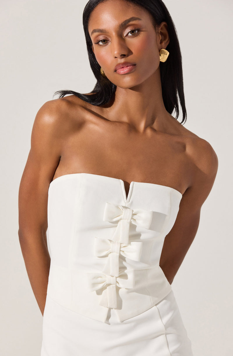 A woman is wearing a strapless white dress with a fitted bodice featuring a small V-shaped slit at the top center. The dress is decorated with a series of three large, neatly tied bows running vertically down the front. She has dark, straight hair and is accessorized with large, gold, semi-circular earrings.