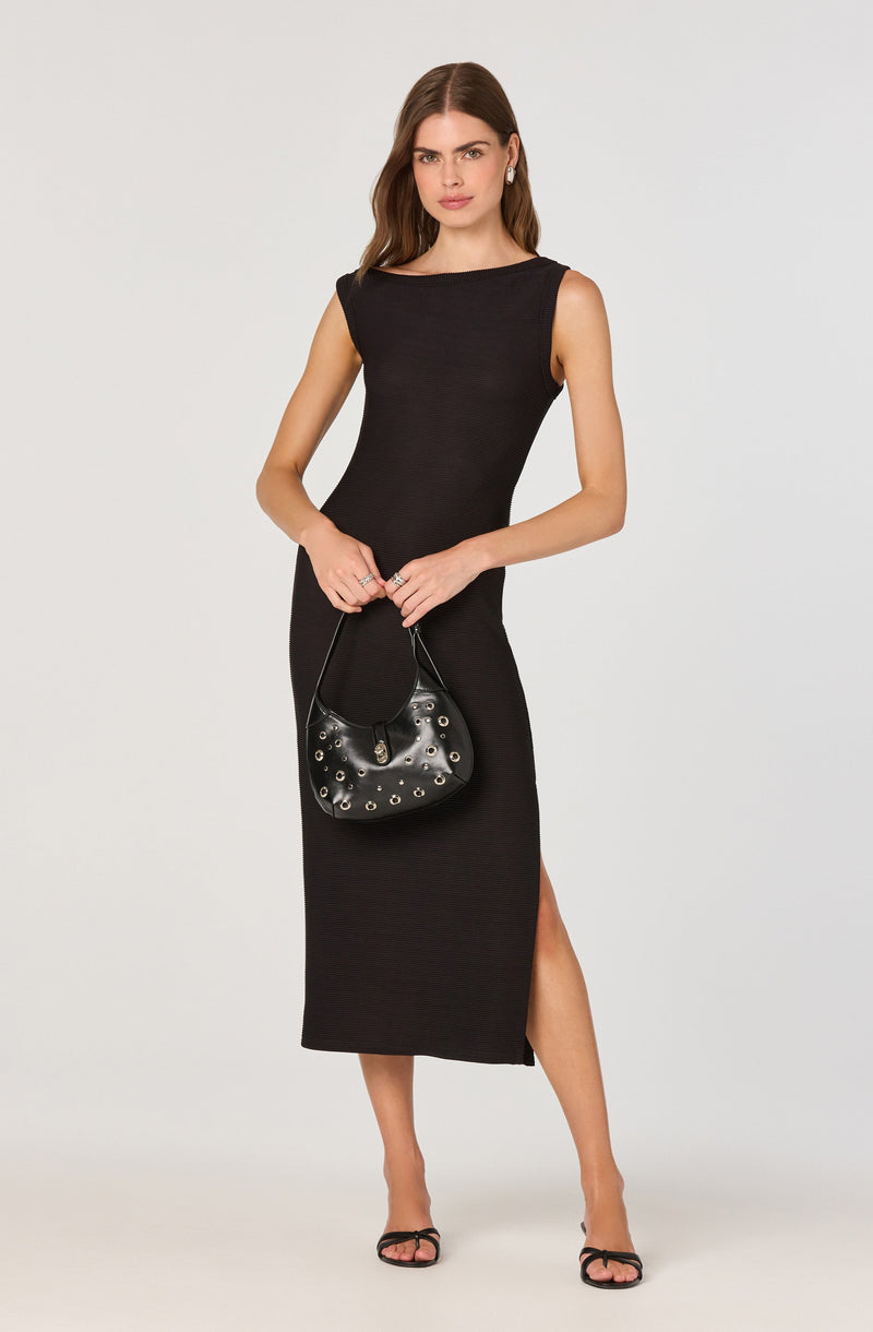 A woman is wearing a sleeveless, black, form-fitting maxi dress with a slit on the side. She is holding a black handbag with metal eyelet details and is wearing black strappy high-heeled sandals.