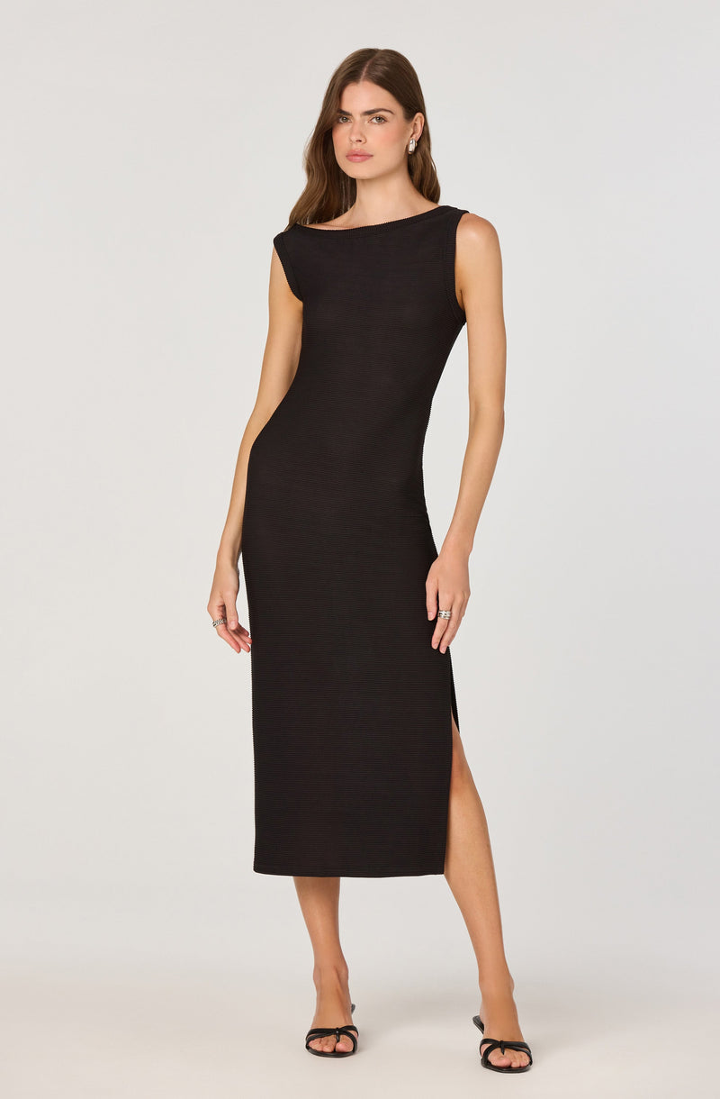 A woman is wearing a sleeveless, black, full-length dress with a high slit on the side. The dress has a boat neckline and a fitted silhouette. She is also wearing black open-toe high-heeled sandals.
