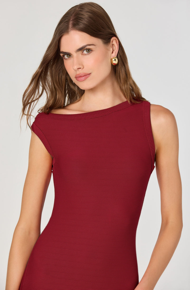 A woman is wearing a sleeveless, form-fitting red dress with a subtle horizontal stripe pattern. The dress has a rounded neckline with the right shoulder slightly exposed and a thicker, ribbed edge along the neckline and armholes. She is accessorized with large, gold hoop earrings.