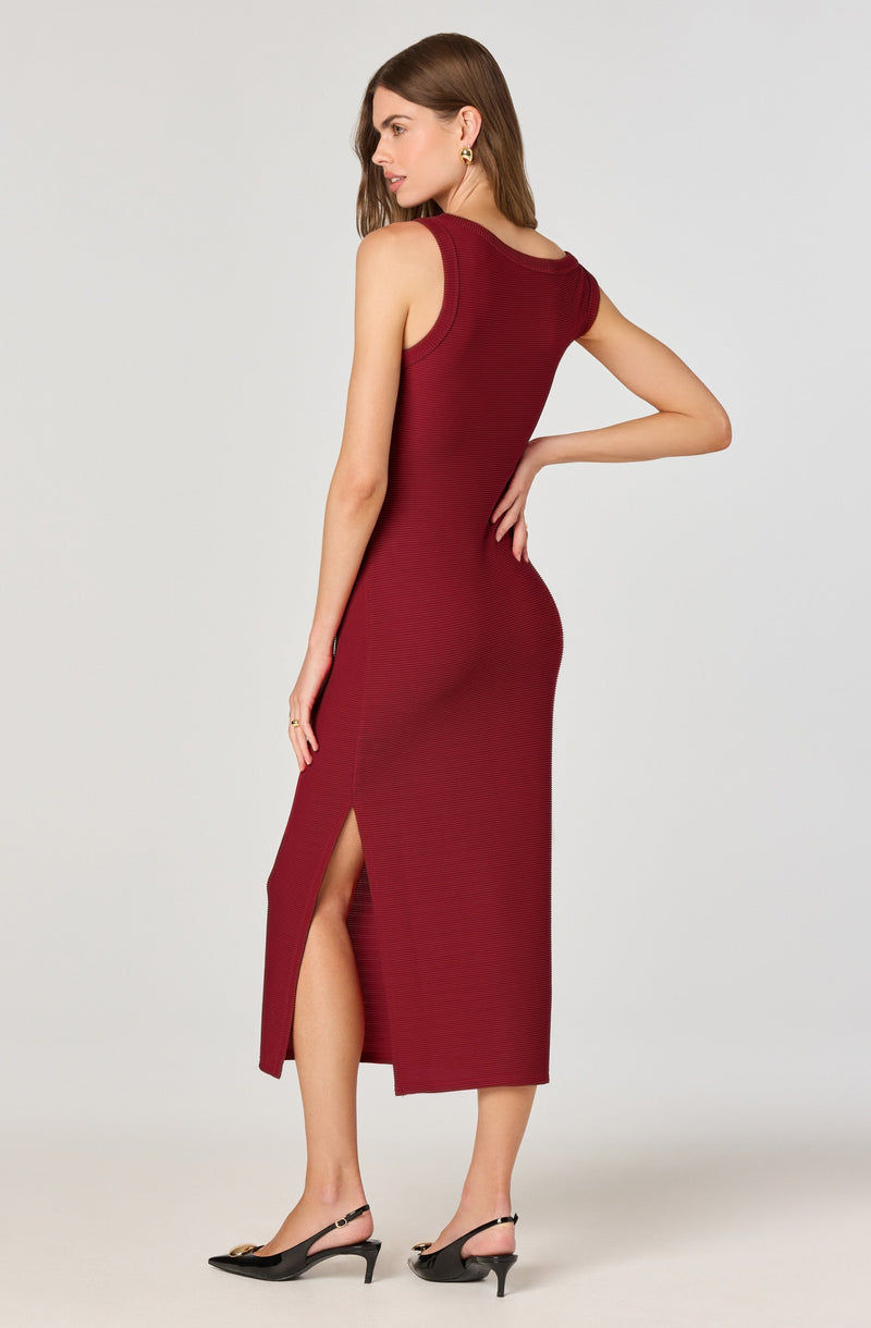 A woman is wearing a sleeveless, ankle-length, deep red dress with a side slit. The dress has a fitted silhouette and features a round neckline. She is pairing the outfit with black high-heeled sandals with ankle straps.