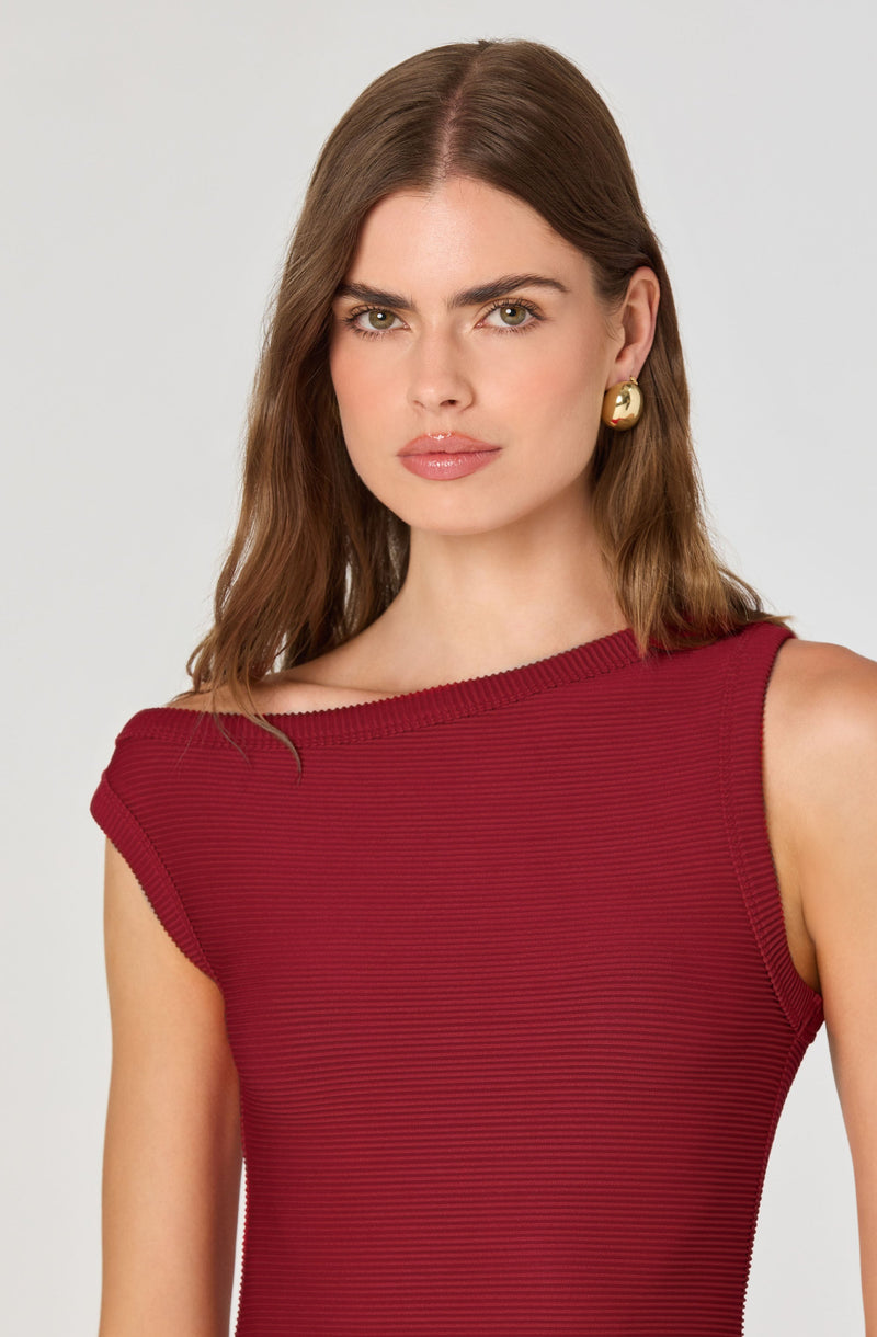 A woman is wearing a sleeveless, form-fitting dress in a rich burgundy color. The dress has a subtle, fine horizontal ribbed texture. She is accessorized with large, gold, round earrings.