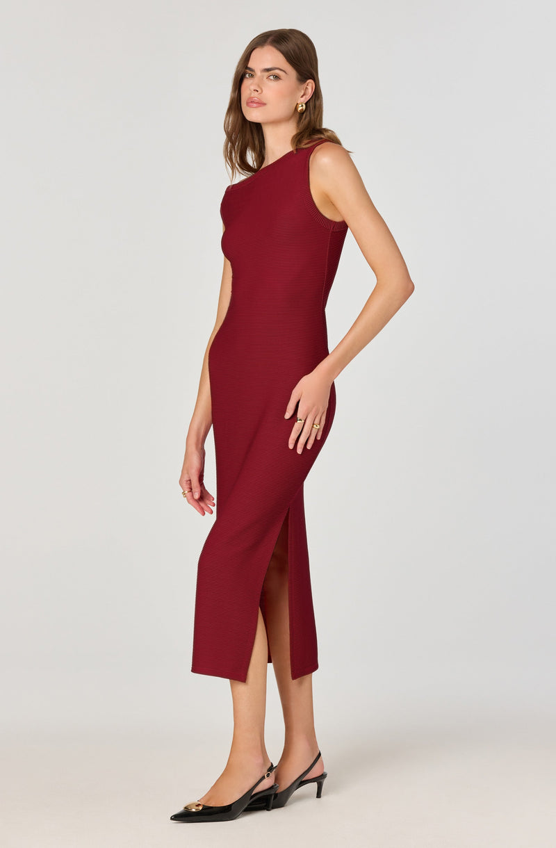 A woman is wearing a sleeveless, form-fitting, midi-length burgundy dress with a subtle texture. The dress features a high slit on the side. She pairs it with black pointed-toe slingback heels with a small heel and gold accents, and gold earrings. She has shoulder-length brown hair and is posing against a plain light gray background.