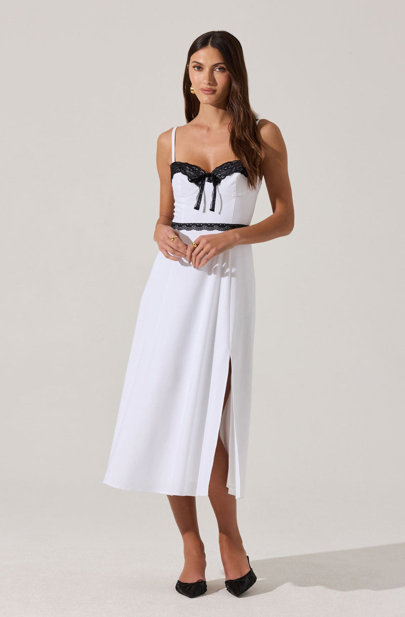 A woman is wearing a white sleeveless dress with spaghetti straps. The bodice features black lace trim along the neckline and a black bow at the center. The waist has a black lace band, and the skirt is long with a front slit, revealing her legs. She is also wearing black pointed-toe heels.