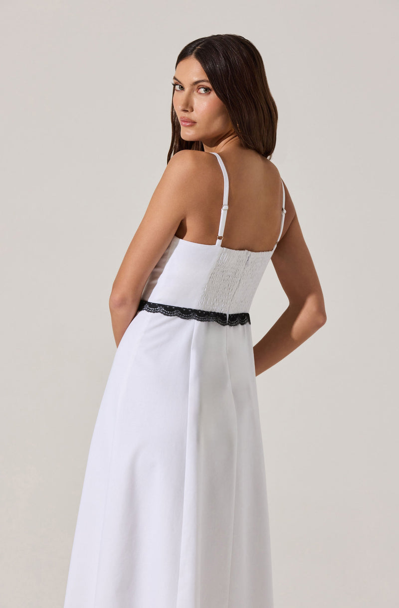 A woman is wearing a long, white dress with thin white shoulder straps. The dress features a fitted shirred bodice and a black lace trim around the waist.