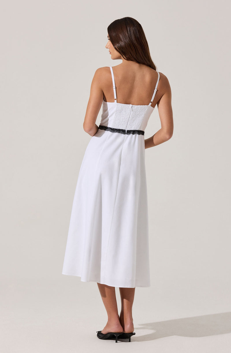 A woman is wearing a white, sleeveless dress with thin adjustable straps. The top part of the dress features smocked detailing and a black lace trim around the waist. The skirt is long, flowing, and reaches mid-calf, with a slight flare. She is also wearing black high-heeled shoes.