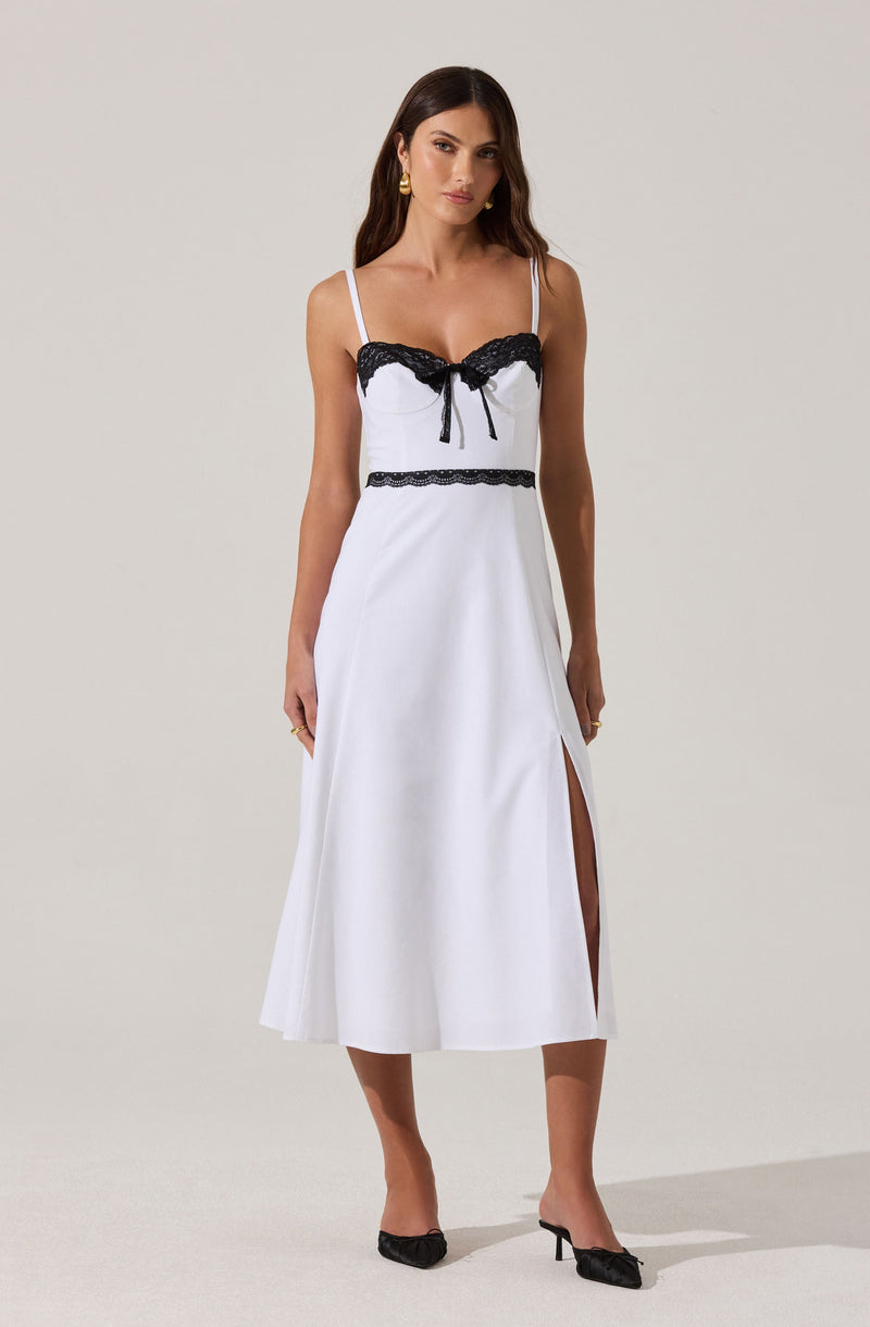 A woman is wearing a sleeveless, white dress with thin spaghetti straps. The dress features black lace trim along the bust line and a small bow in the center. There is a horizontal black lace band at the waist and a slit on the left side of the skirt. The woman is also wearing black open-toe high heels with bows.