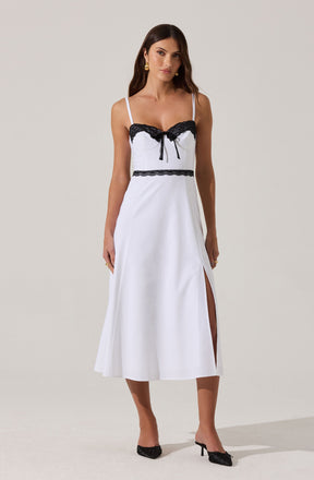 A woman is wearing a sleeveless, white dress with thin spaghetti straps. The dress features black lace trim along the bust line and a small bow in the center. There is a horizontal black lace band at the waist and a slit on the left side of the skirt. The woman is also wearing black open-toe high heels with bows.