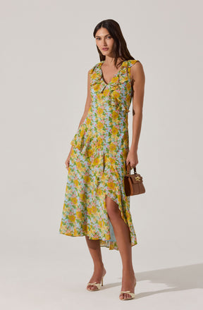 A woman is wearing a sleeveless, ankle-length dress with a floral pattern in shades of yellow, pink, green, and white. The dress features ruffled details around the armholes and neckline, and has a slit on one side. She is paired with open-toed high-heeled shoes and is holding a small brown handbag.