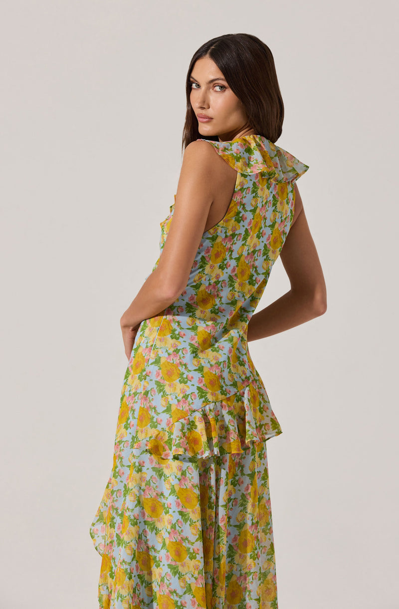 A woman is wearing a sleeveless, floral dress with a ruffled detail on the shoulders and a ruffle at the waistline. The dress features a colorful pattern of yellow, pink, and green flowers on a light background. The fabric appears lightweight and flowy, suitable for warm weather.