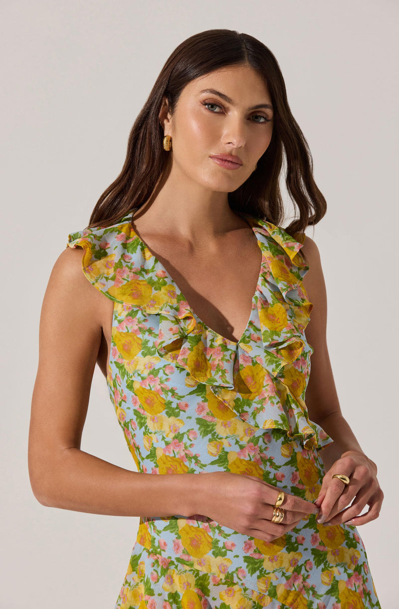 A woman is wearing a sleeveless dress with a floral pattern featuring yellow flowers, pink accents, and green leaves on a light background. The dress has a V-neckline with ruffled detailing along the edges that extends over the shoulders.