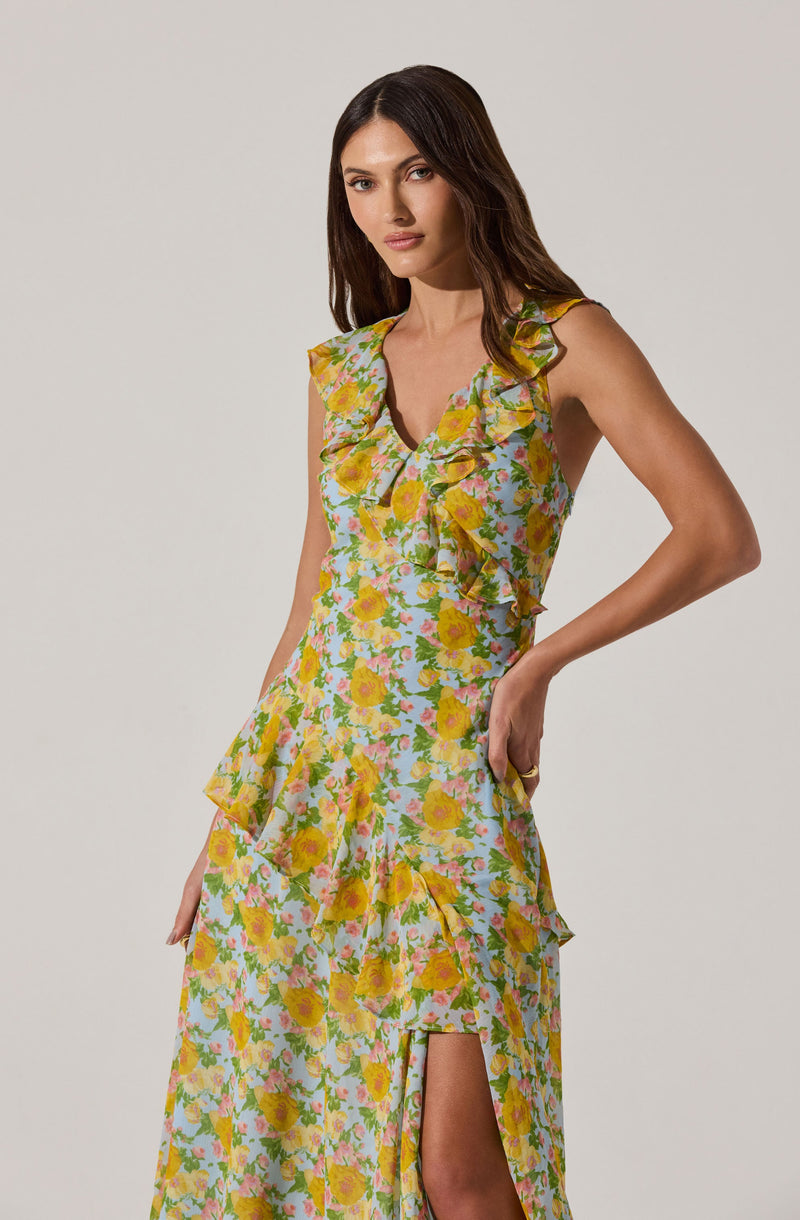A woman is wearing a sleeveless, floral maxi dress with a light pastel-colored background. The dress features a ruffled neckline, ruffles along the waistline, and a front slit that reveals part of her leg. The floral pattern consists of yellow, pink, and green flowers.