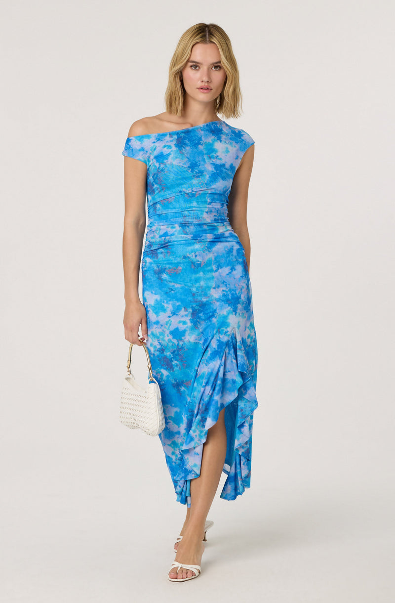 A woman is wearing a blue tie-dye dress with an asymmetrical off-the-shoulder neckline, ruched details around the waist, and a ruffled, high-low hemline. She is holding a small white woven handbag and wearing white open-toe high heels.