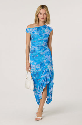 A woman is wearing a blue tie-dye dress with an asymmetrical off-the-shoulder neckline, ruched details around the waist, and a ruffled, high-low hemline. She is holding a small white woven handbag and wearing white open-toe high heels.