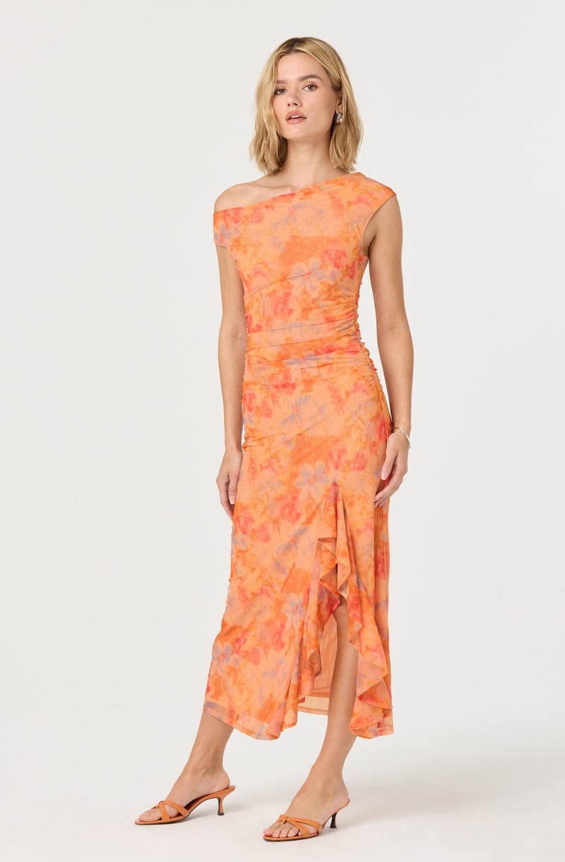 A woman is wearing a sleeveless, off-the-shoulder dress with a tie-dye pattern in shades of orange, pink, and purple. The dress has a fitted silhouette with ruched details and a ruffled, asymmetrical hemline that reveals a slit in the front. She completes her outfit with matching orange strappy high-heeled sandals.