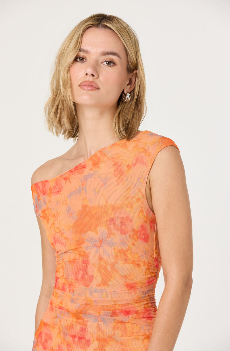 A woman is wearing a sleeveless, one-shoulder dress with a fitted silhouette. The dress features a vibrant, abstract floral pattern in shades of orange, pink, and purple, with a slightly ruched texture throughout the fabric.
