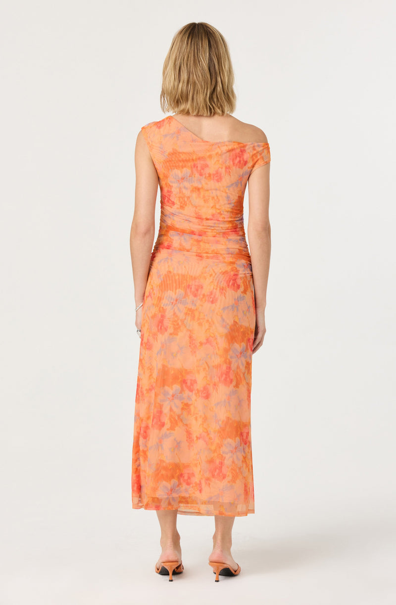 A woman is wearing a sleeveless, off-the-shoulder maxi dress with a vibrant, abstract orange, pink, and purple floral pattern. The dress has a fitted waist and flows down to her ankles. She is also wearing nude-colored heeled sandals.