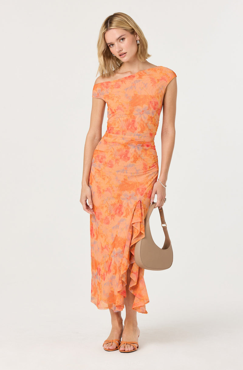 A woman is wearing a sleeveless, floor-length dress with an off-the-shoulder neckline. The dress features a tie-dye pattern in shades of orange, pink, and purple, and has a ruffle detail along one side of the hem. She is holding a beige, crescent-shaped handbag and wearing strappy sandals that match the color of the dress.