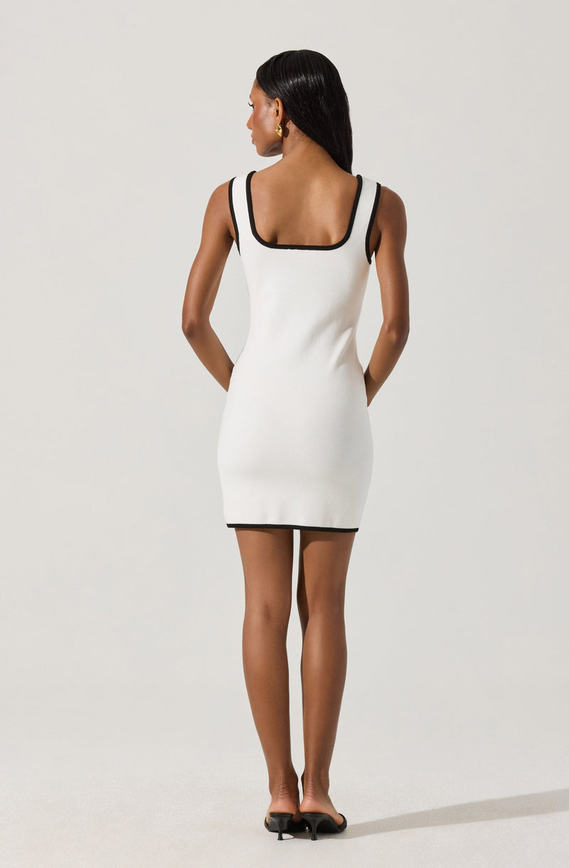 A woman is wearing a sleeveless, fitted white dress with black trim along the neckline, armholes, and hemline. She has long black hair and is accessorized with gold hoop earrings. She is standing against a plain, light-colored background.