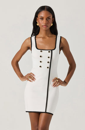 A woman is wearing a fitted sleeveless dress that is primarily white with black trim along the neckline, armholes, and a vertical line running down the front. The dress features decorative black buttons arranged in two vertical rows on the front. The dress has a square neckline and a straight hemline that ends above the knees. She accessorizes with gold earrings and rings.