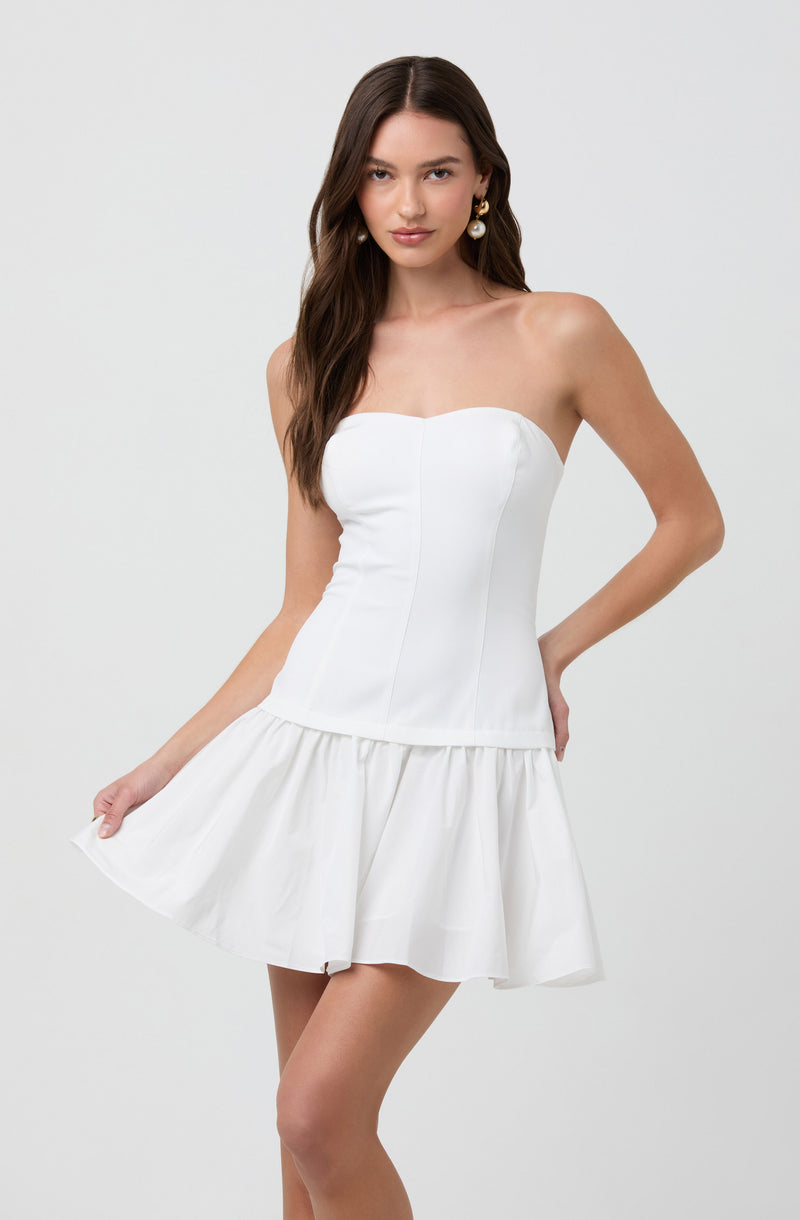 A young woman is wearing a strapless white dress with a fitted bodice, featuring vertical seams and a ruffled, flared skirt that ends above the knee. She has long, wavy brown hair and is accessorized with large, round, pearl earrings.
