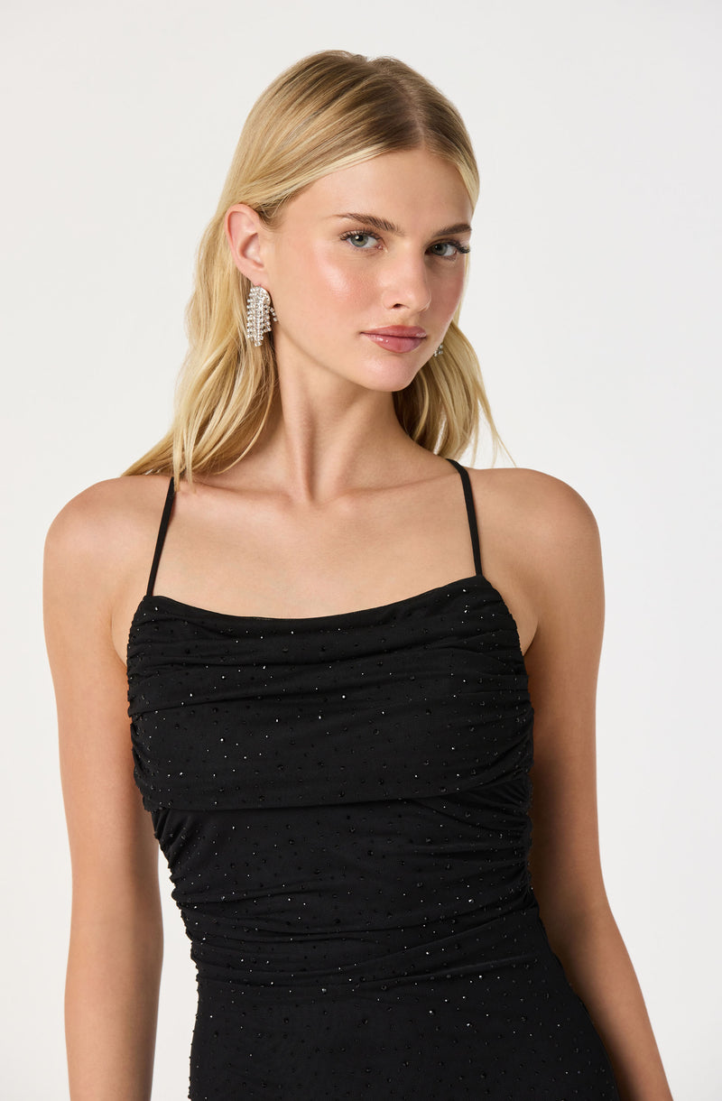 A woman is wearing a black spaghetti strap dress that is ruched along the bodice and adorned with small, sparkly embellishments.