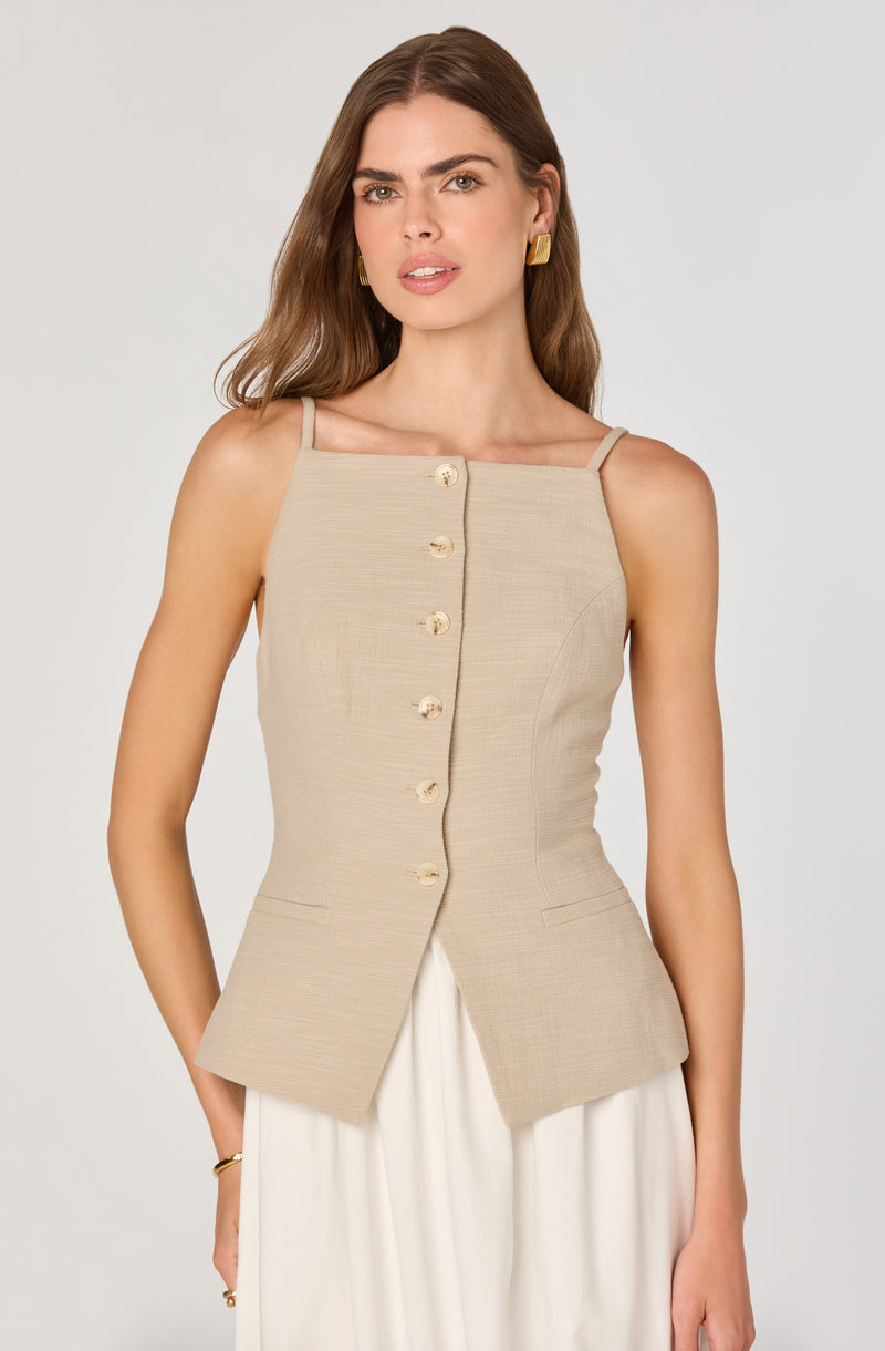 A woman is wearing a sleeveless beige vest with thin spaghetti shoulder straps. The vest features a row of decorative buttons down the front and has a fitted silhouette with a slight flare at the hips. It is paired with a white skirt.