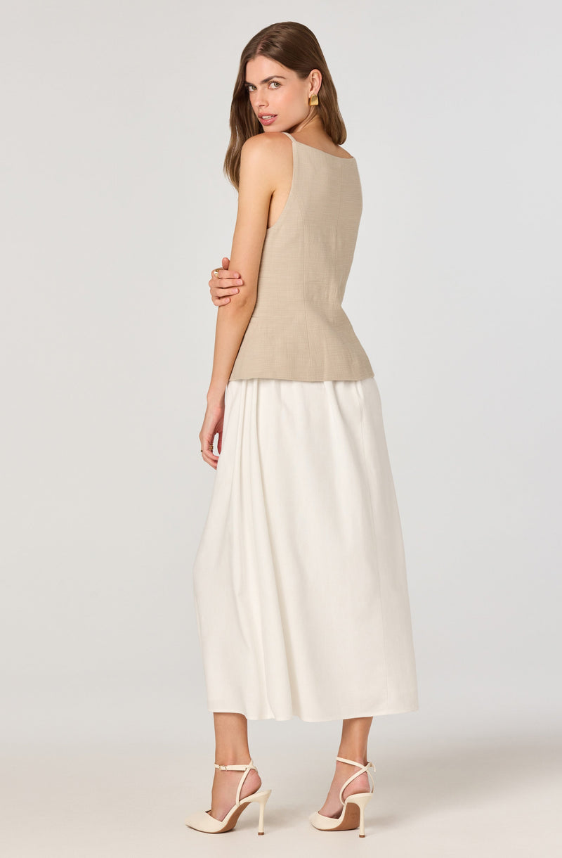 A woman is wearing a sleeveless beige top with a fitted bodice and a subtle textured pattern. She pairs it with a long, flowing white skirt that reaches her ankles. She completes her outfit with cream-colored high-heeled sandals that have ankle straps.