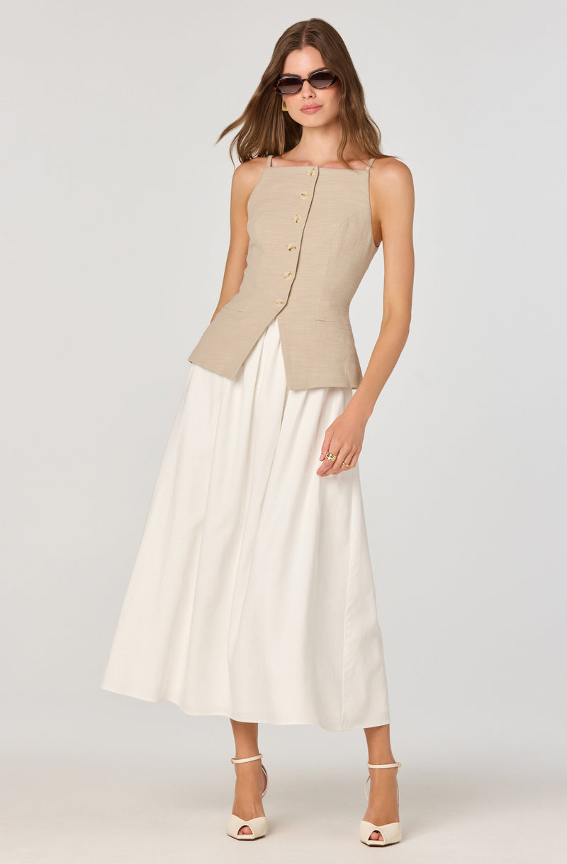 A woman is wearing a sleeveless, beige vest with a row of buttons down the front, paired with a long, flowing white skirt. She has on white high-heeled sandals and is accessorized with sunglasses.