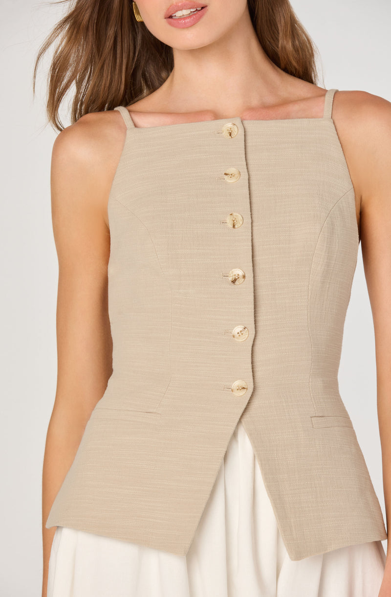 A woman is wearing a sleeveless beige vest with thin shoulder straps. The vest features a front row of shiny, gold-colored buttons that run down the center. The fabric appears textured with subtle lines, and the vest has a tailored fit with curved seam details. She is also wearing a white skirt or pants underneath.