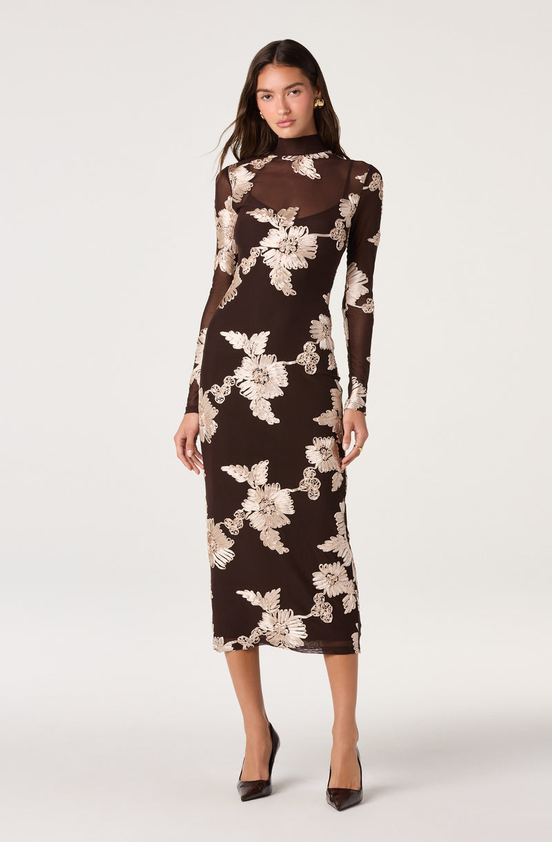 Woman wearing a long black dress with beige floral embroidery and sheer long sleeves.