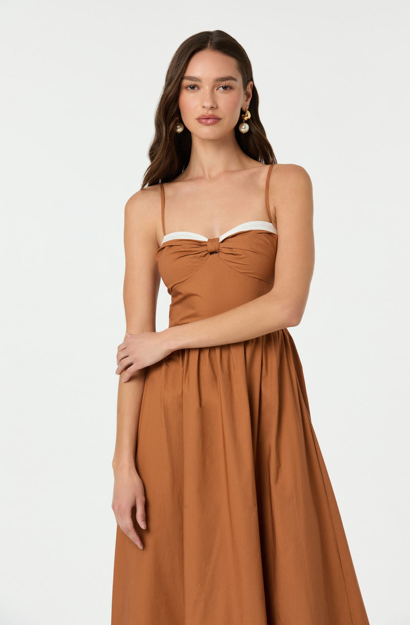 A young woman is wearing a burnt orange dress with spaghetti straps. The top of the dress features a white band detail across the bust, with a small knot in the center. The skirt is flowy and gathered at the waist, creating a full, elegant silhouette.