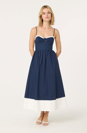 A woman is wearing a sleeveless, navy blue maxi dress with thin spaghetti straps. The dress features white trim along the top edge and a small bow detail at the center of the chest. The skirt is gathered at the waist and has a wide white band at the hem. She is also wearing white open-toe heels.