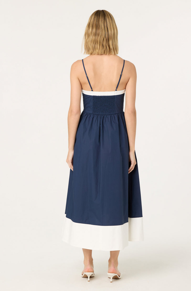 The dress features thin spaghetti straps over the shoulders, a fitted bodice with a smocked waistband, and a full, midi-length skirt. The dress has a color block design with navy blue on the upper part and the majority of the skirt, and a white band near the hem. The fabric appears lightweight and flowy. The model is wearing white heeled sandals.