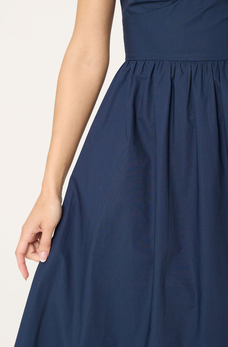 A person is wearing a navy blue dress with a fitted bodice and a high waistband. The skirt is gathered and flows outward, creating a full, A-line silhouette. The fabric appears to be smooth with a slight sheen. The dress has a sleeveless design, and the person's hand is resting gently at their side.