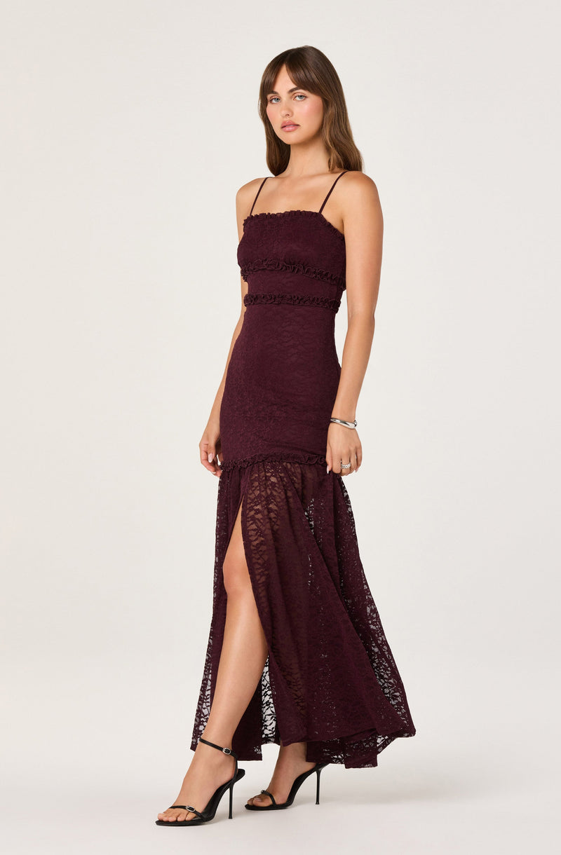 A woman is wearing a floor-length, sleeveless dress with thin spaghetti straps. The dress is made of a dark burgundy lace fabric with horizontal lace bands on the bodice and waist. The skirt features a high slit on one side, revealing her leg and the matching lace lining underneath. She is also wearing black high-heeled sandals and a simple bracelet on her left wrist.