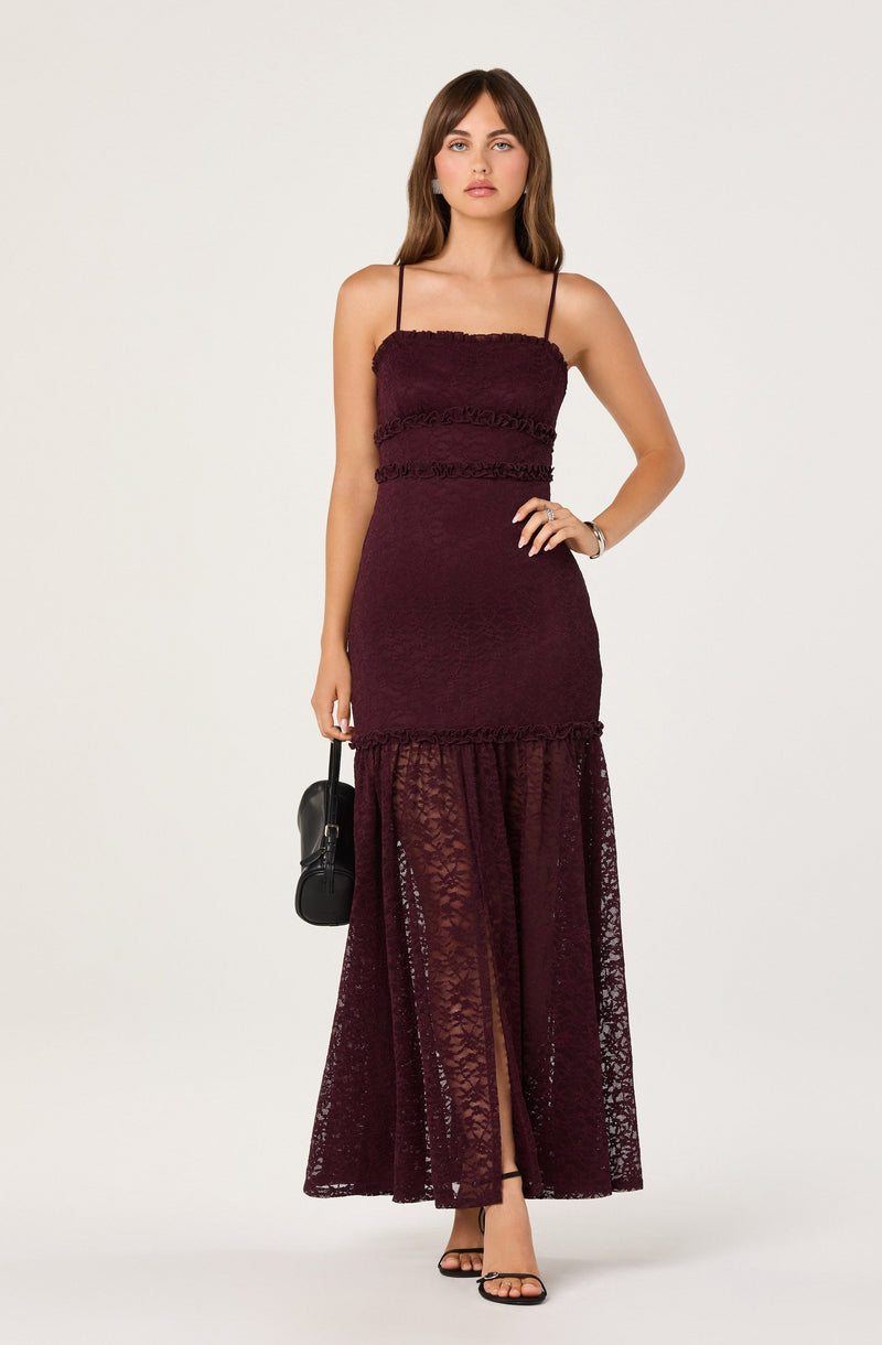 The model is wearing a sleeveless, burgundy dress with thin spaghetti straps. The dress has a fitted bodice with textured horizontal stripes and a sheer, lace overlay from the mid-thigh down to the floor, showing a slit at the front. The lace overlay features floral patterns and extends to the ground.