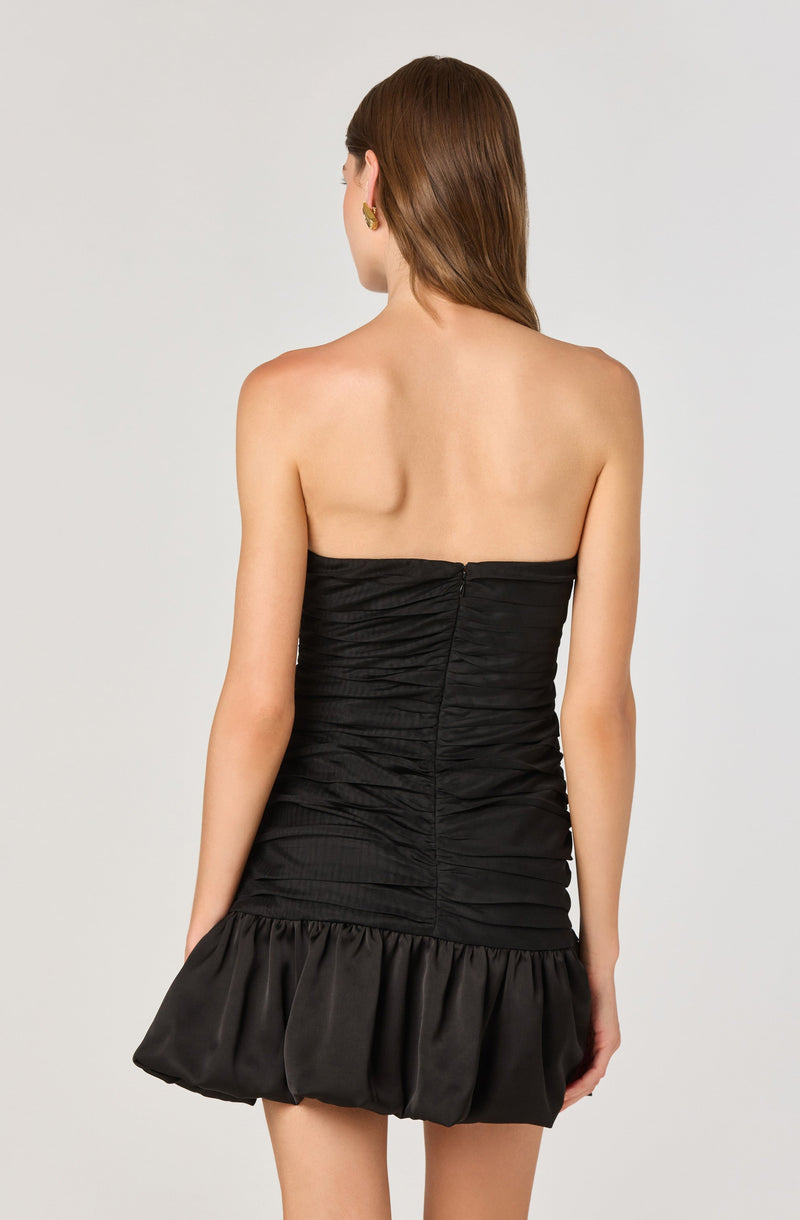 A woman is wearing a strapless black dress with a ruched bodice and a ruffled, gathered satin skirt at the hem. The dress is fitted and short, ending above the knees. She has earrings on and her brown hair is styled straight, falling over her shoulders.