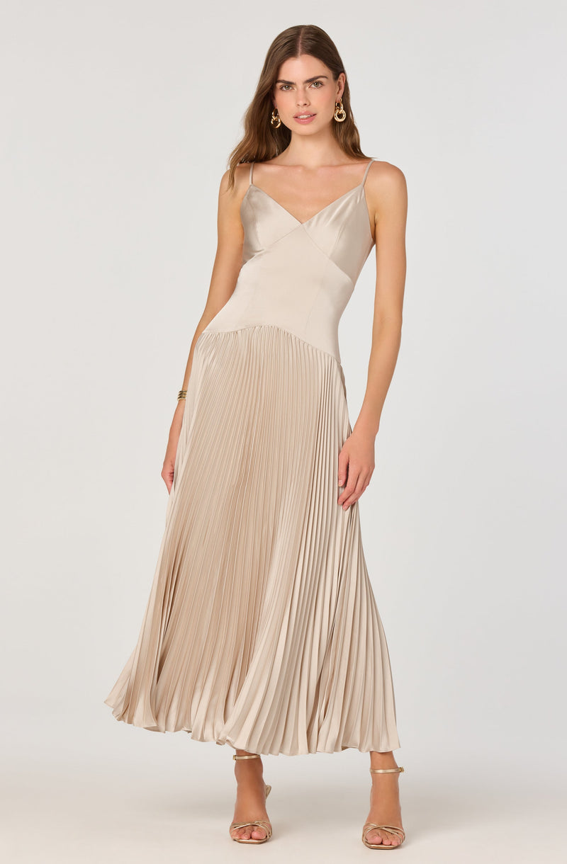 A woman is wearing a sleeveless, satin dress with thin spaghetti straps. The top part of the dress is smooth and fitted, with a V-neckline. The skirt is pleated and flows down to the floor, creating a long and elegant silhouette. She is also wearing high-heeled sandals with ankle straps and has large hoop earrings as accessories.