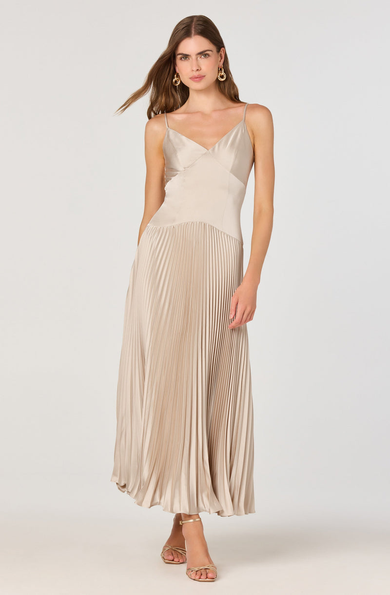 A woman is wearing a sleeveless, beige-colored dress with thin spaghetti straps. The top part of the dress has a smooth, satin-like finish with a V-neckline, while the skirt features a pleated design that creates a flowing and elegant silhouette. The dress is floor-length, and she is wearing open-toe heels.
