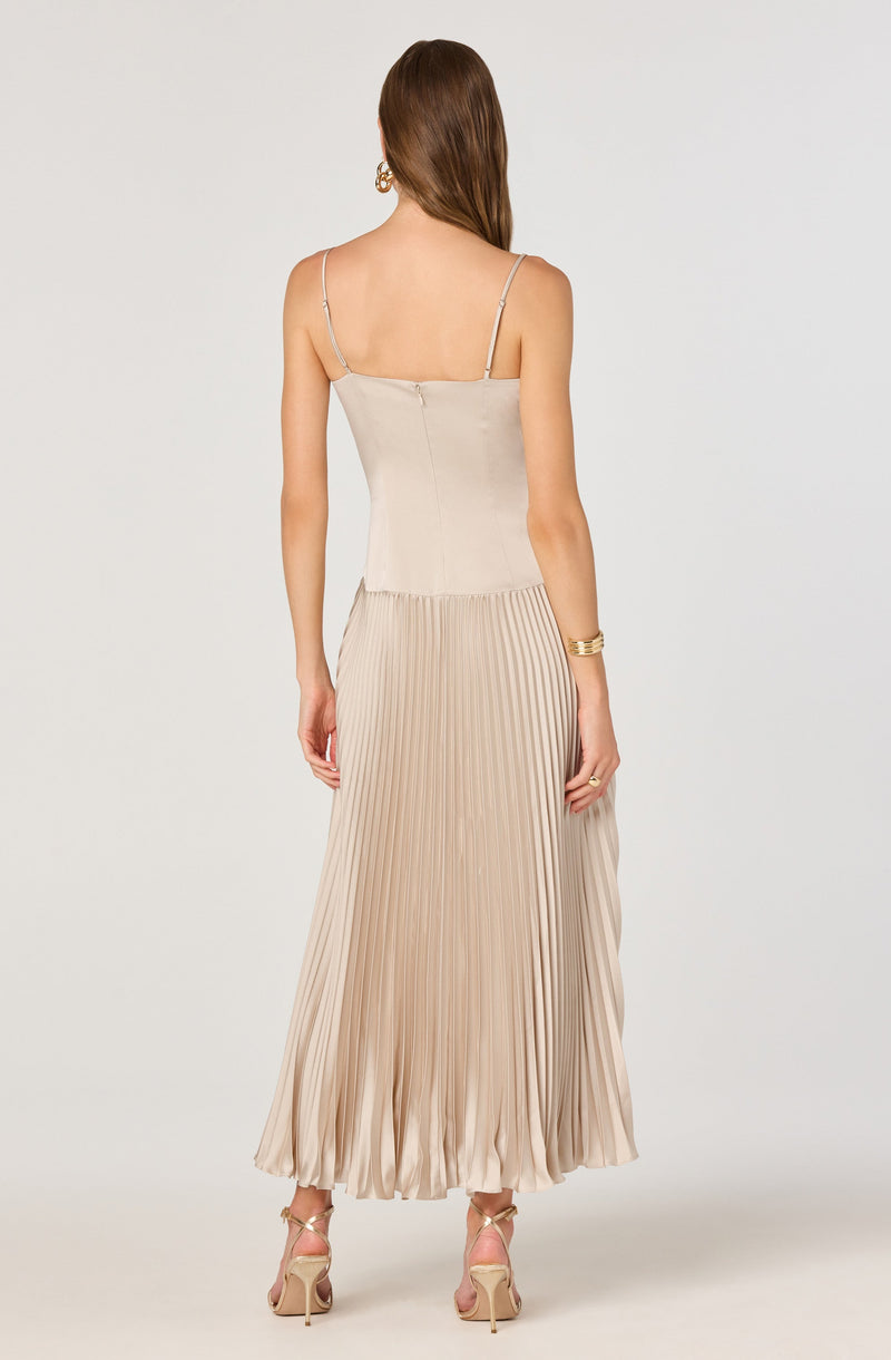 The woman is wearing a beige sleeveless top with thin spaghetti straps and a high-waisted, pleated, satin maxi skirt in a matching shade. She also has on high-heeled sandals with ankle straps, large hoop earrings, and a gold bangle bracelet on her left wrist.
