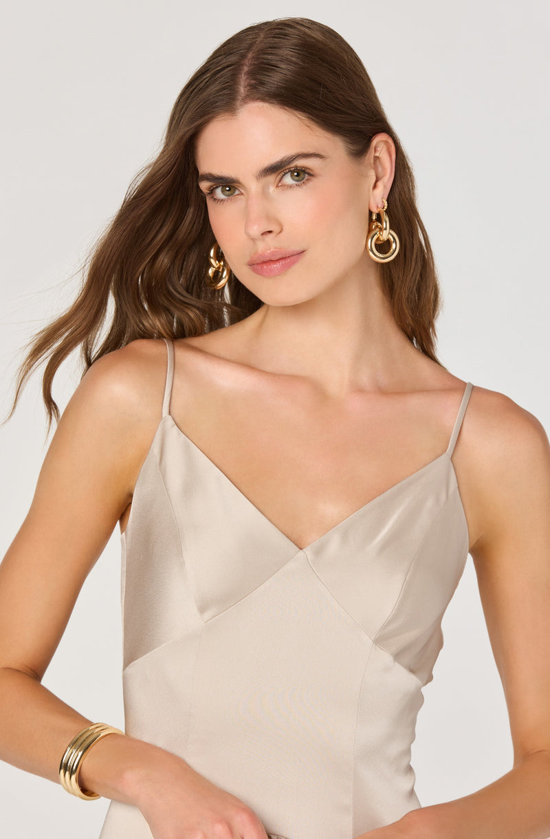 A woman is wearing a sleeveless, satin-like dress with thin spaghetti straps. The dress has a V-neckline and features a color-block design with panels in a light, neutral shade. She accessorizes with large, gold hoop earrings and gold bangles on her wrist.
