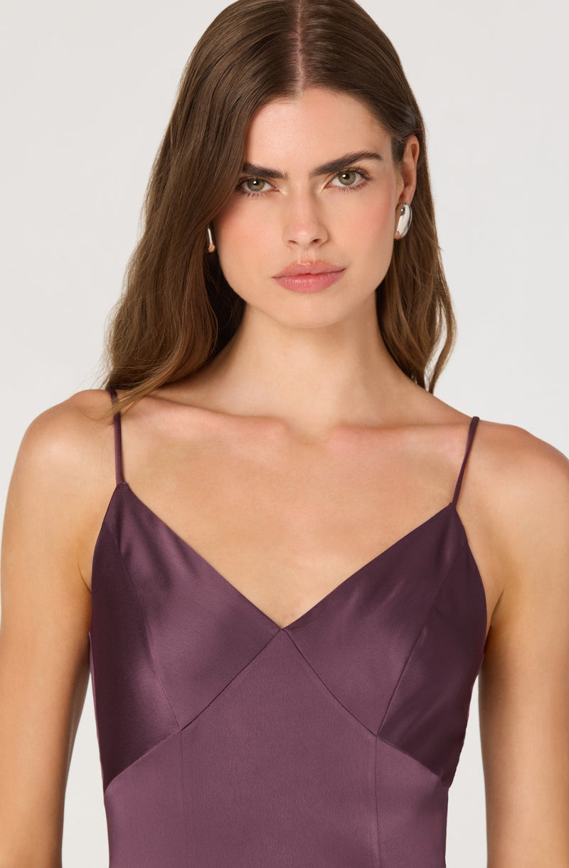 A woman is wearing a sleeveless dress with thin spaghetti straps. The dress has a satin finish and features a color-block design with shades of dark purple and plum, creating a diagonal pattern across the bodice.