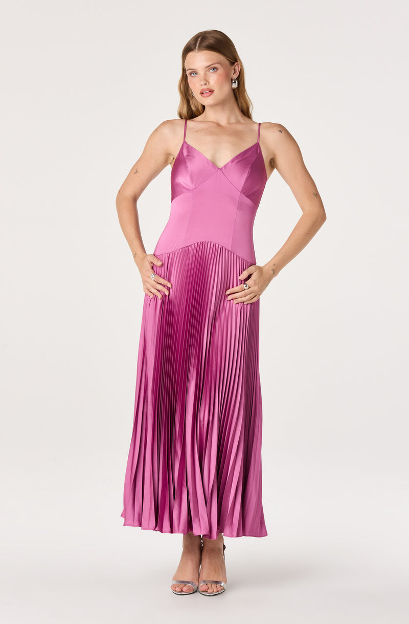 Woman in a pink satin and pleated maxi dress with spaghetti straps, standing against a white background.