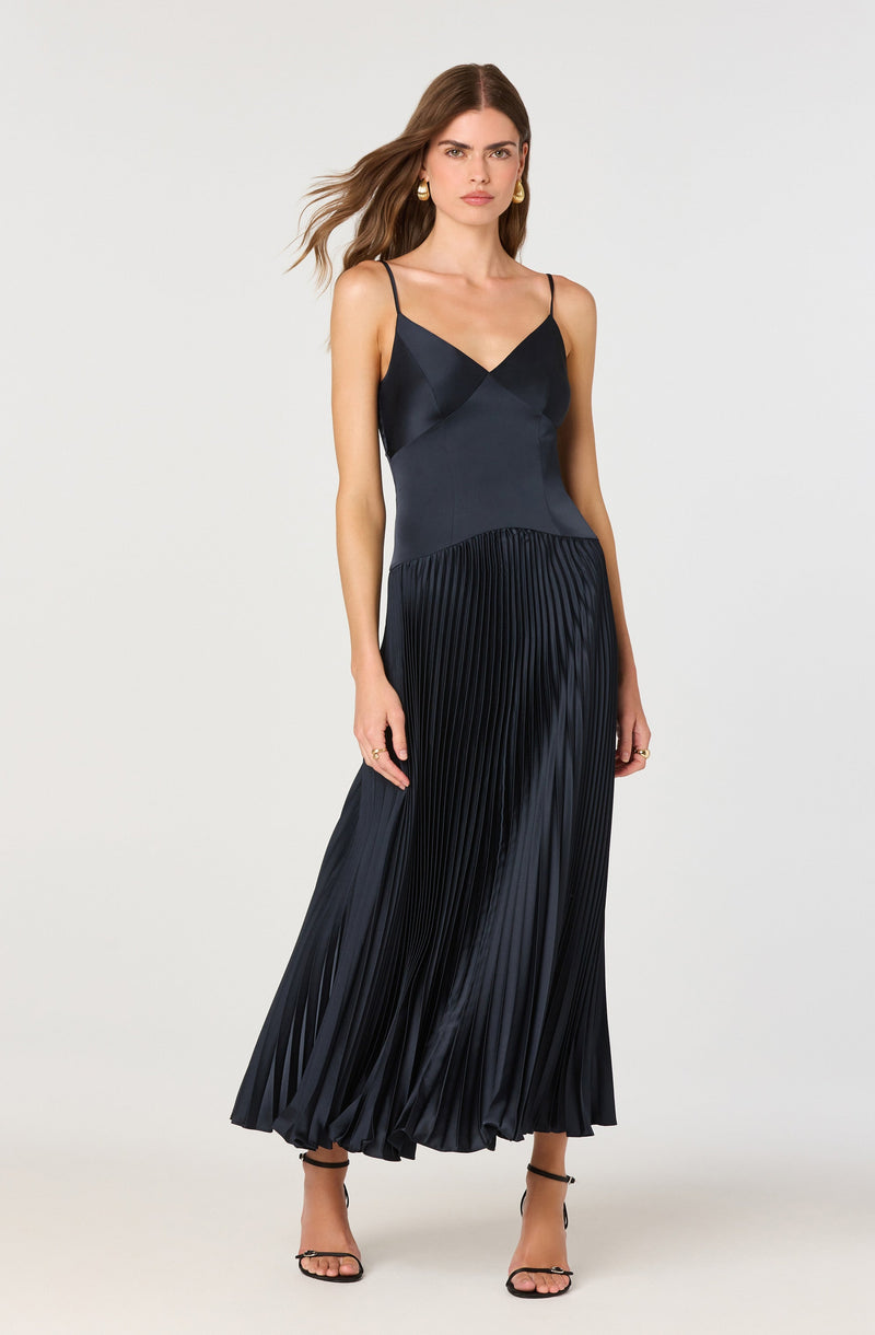 A woman is wearing a long, elegant navy blue dress with thin spaghetti straps. The top of the dress features a smooth, satin-like fabric with a subtle V neckline. The skirt has a pleated design that creates a flowing, voluminous effect, with the pleats adding texture and movement to the dress.